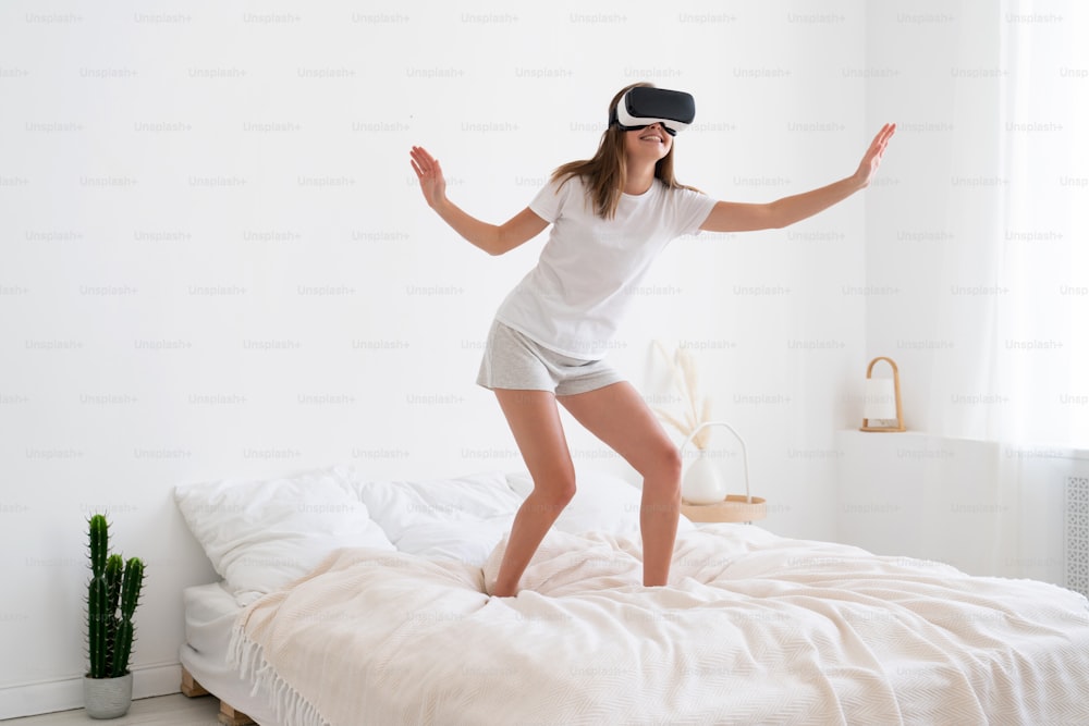 Young woman having fan walking on her bed, enjoying virtual reality in VR headset, moving arms for balancing, smiling and laughing happily