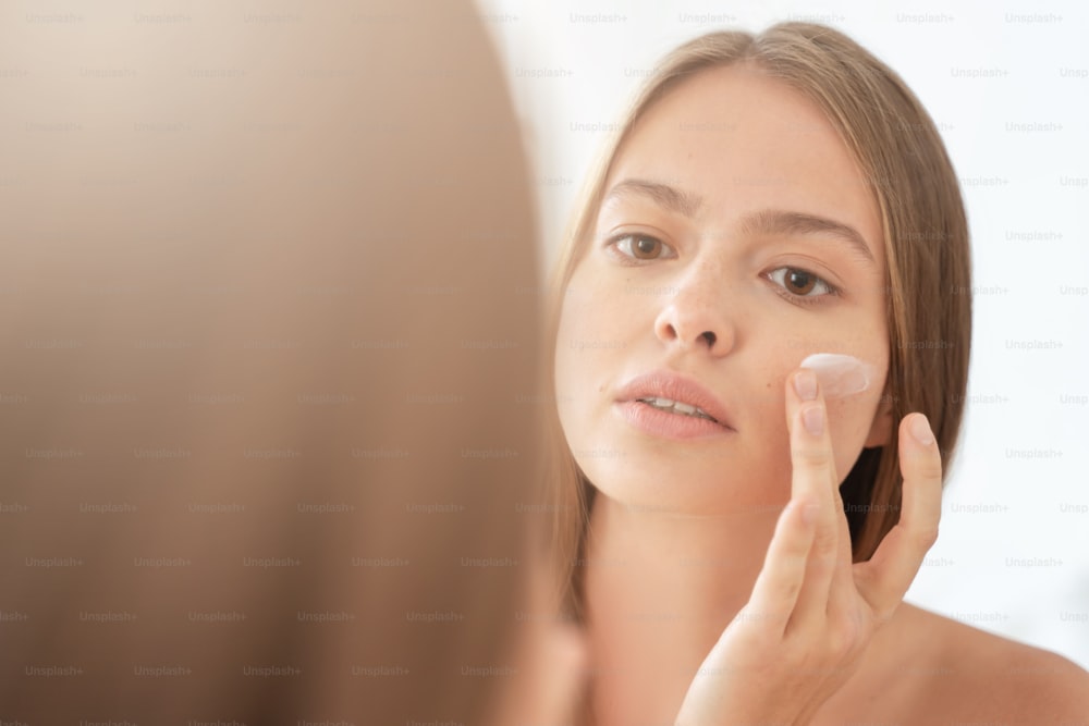 Headshot di giovane bella donna che applica la crema sul suo viso, guardando il suo riflesso nello specchio, prendendosi cura di se stessa, idratando la pelle al mattino