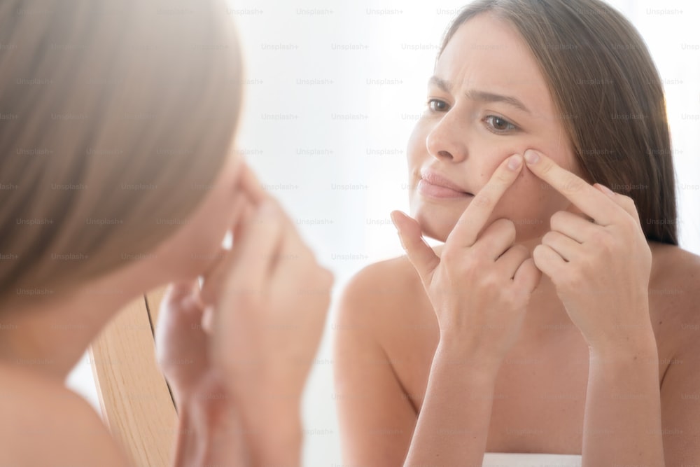 Junge Frau, die in den Badezimmerspiegel schaut, verzweifelt versucht, Pickel auf ihre Wange zu drücken, sich wegen Hautproblemen geärgert