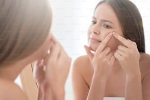 バスルームの鏡を見て、頬のにきびを必死に絞ろうとし、肌の問題のためにイライラしている若い女性