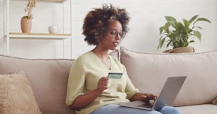 Giovane donna afroamericana che paga online con carta di credito e laptop, acquistando prodotti sul sito di e-commerce, seduta sul divano a casa