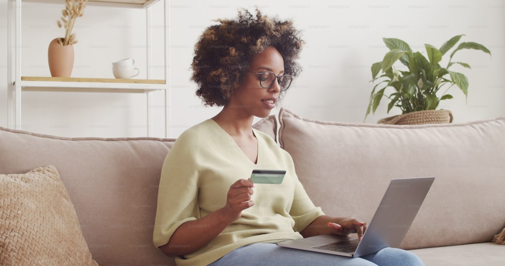 Young african american woman paying online with credit card and laptop, buying products on ecommerce website, sitting on couch at home