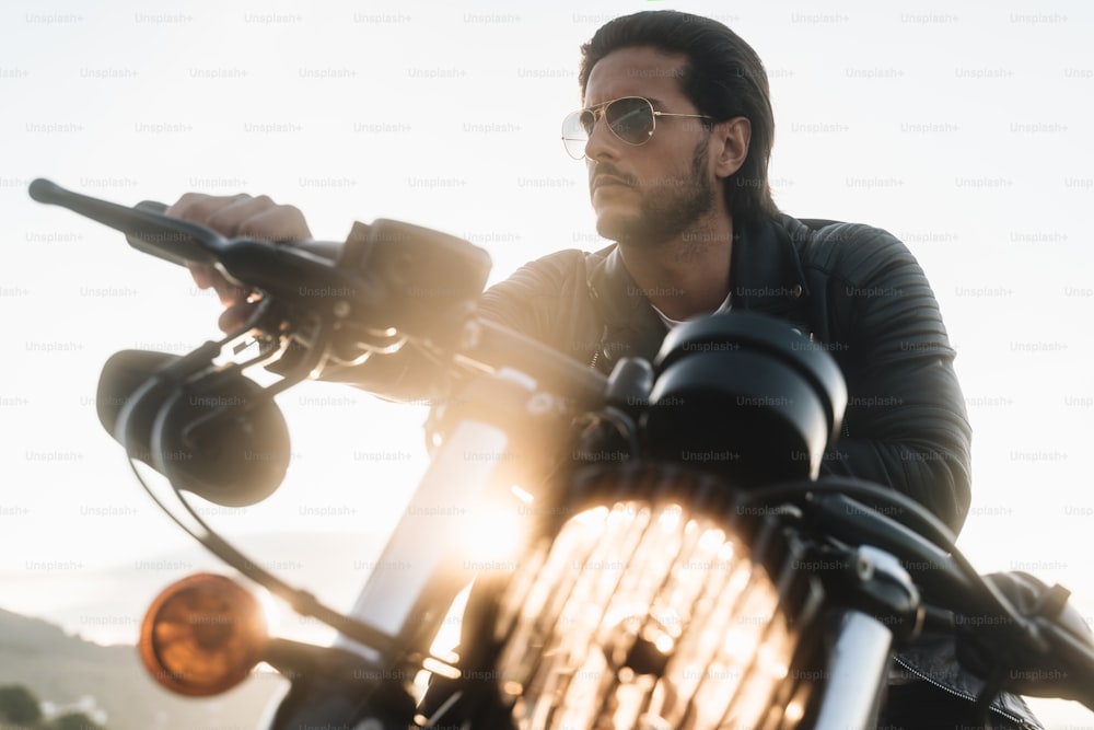 Handsome biker looking away, sitting on his vintage motor bike, wearing leather jacket and stylish sunglasses