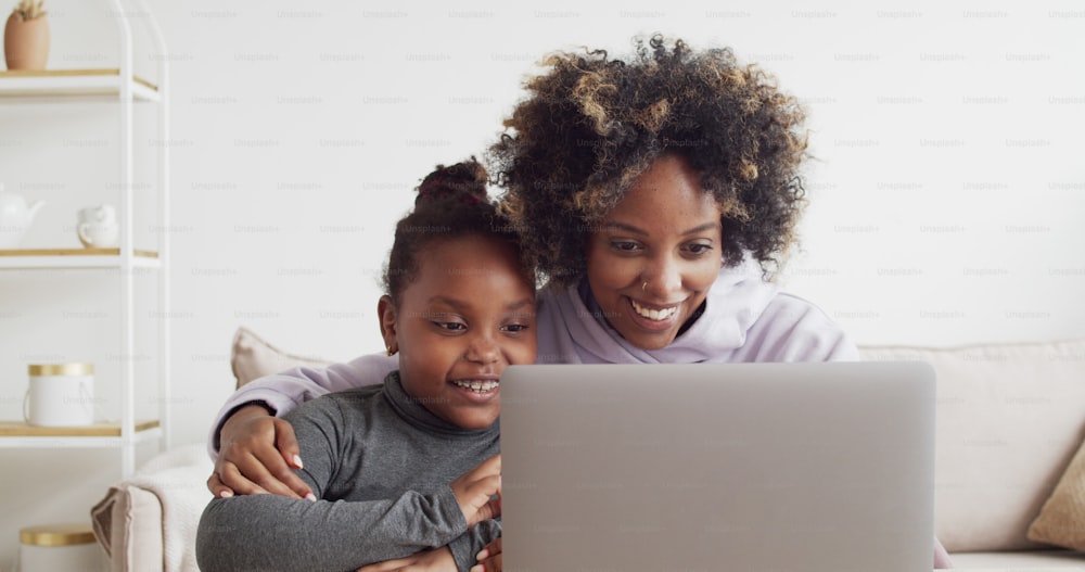 Lächelnde afrikanische Mutter und kleines Mädchen schauen sich zu Hause einen lustigen Film auf dem Laptop an