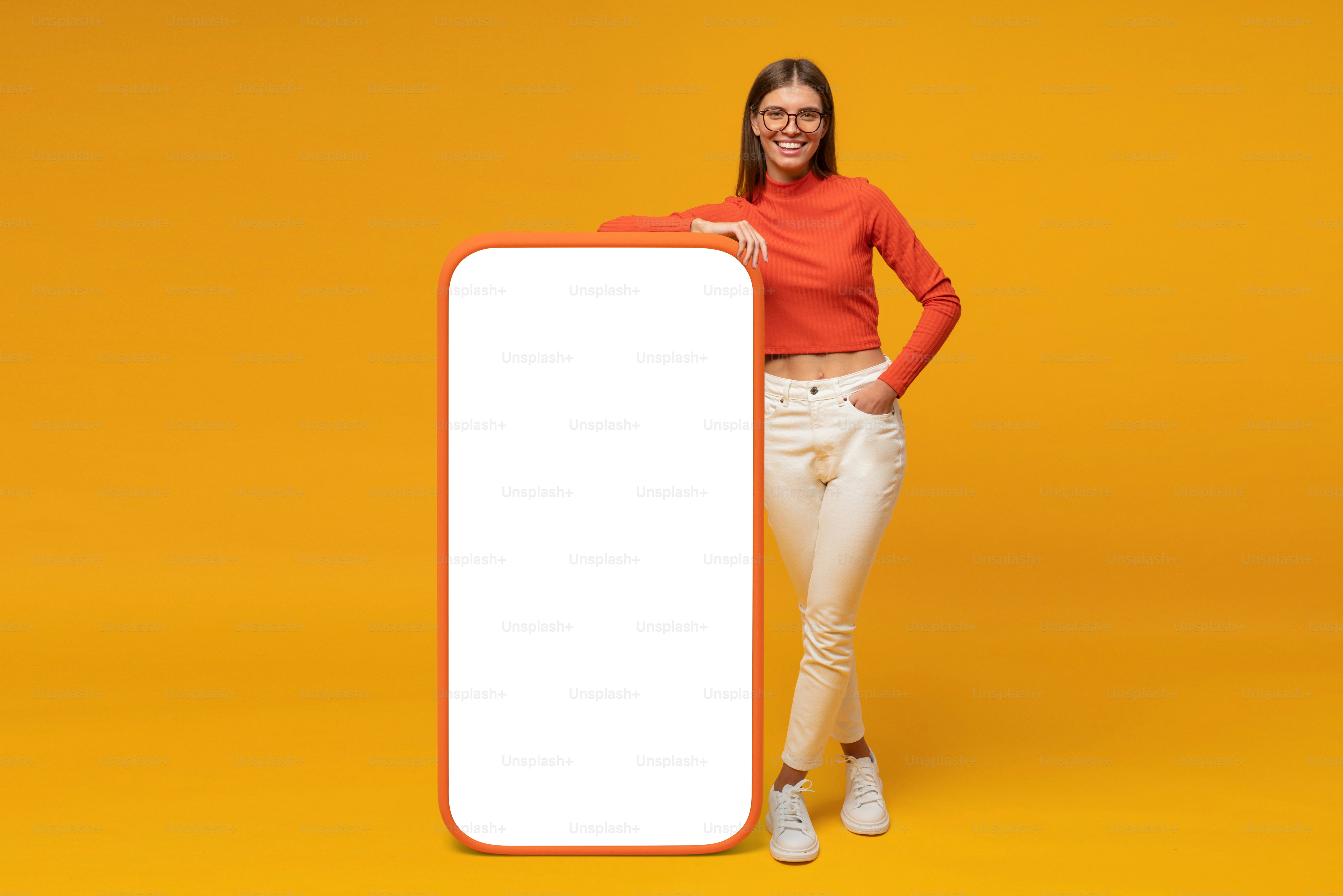 Happy young woman standing and showing blank screen of huge phone, mockup for your app, isolated on yellow background