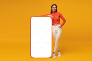 Glückliche junge Frau, die steht und einen leeren Bildschirm eines riesigen Telefons zeigt, Modell für Ihre App, isoliert auf gelbem Hintergrund