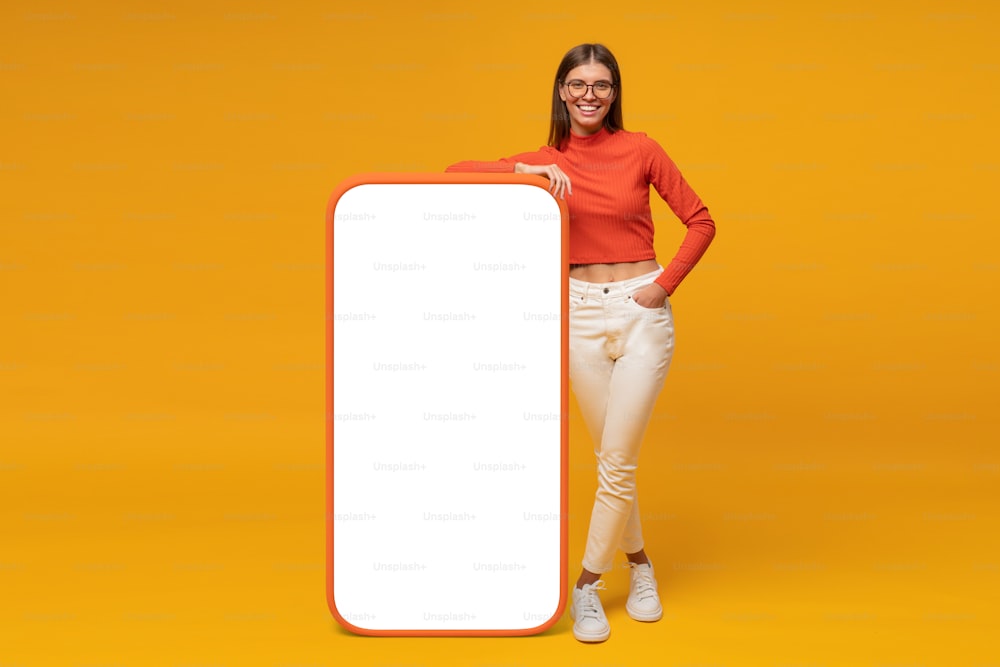 Giovane donna felice in piedi e che mostra lo schermo vuoto del telefono enorme, mockup per la tua app, isolato su sfondo giallo