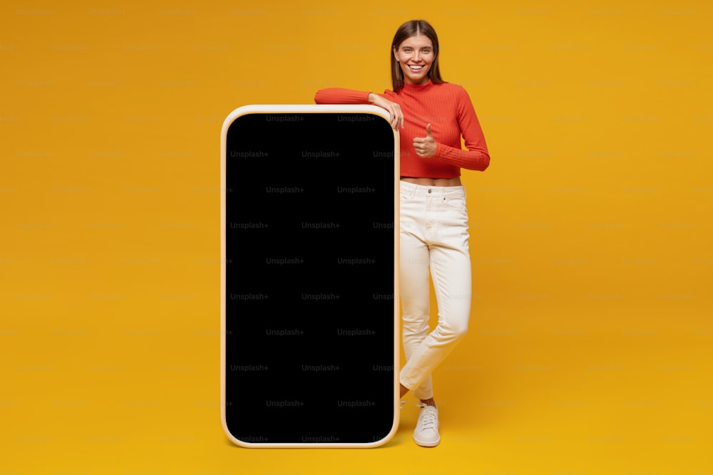 Junge Frau steht und zeigt einen leeren Bildschirm eines riesigen Telefons, Modell für Ihre App, isoliert auf gelbem Hintergrund