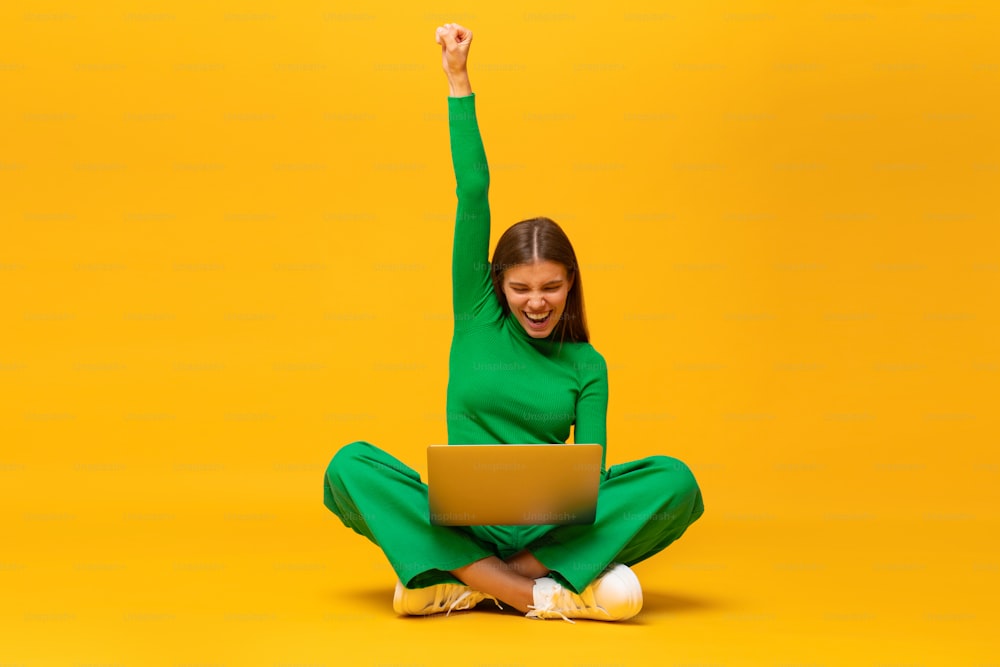 Winner. Excited happy student teen girl sitting on floor with laptop, raising one hand in the air if saying yes, isolated on yellow background