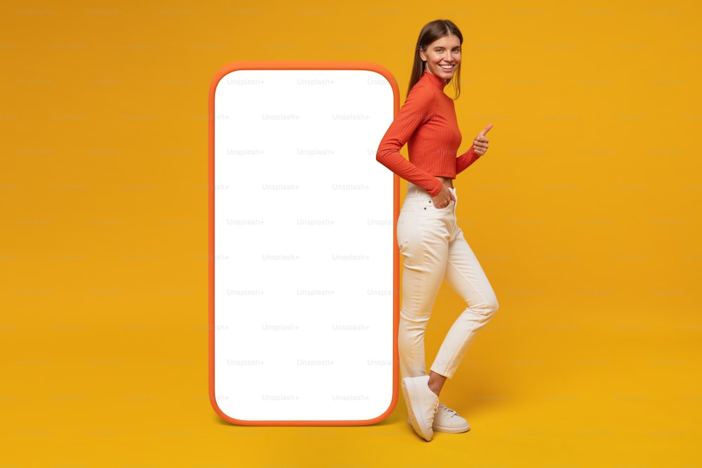 Mujer de pie y mostrando la pantalla en blanco de un teléfono enorme y un gesto de pulgar hacia arriba, maqueta para su aplicación, aislada sobre fondo amarillo