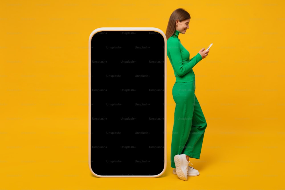 黄色い背景にアプリ用の巨大な電話モックアップの近くに立つ若い笑顔の女性