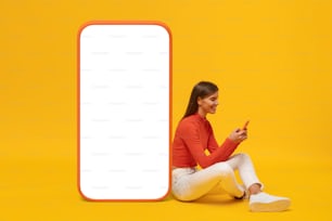 Mujer joven sentada en el suelo cerca de una enorme maqueta de teléfono para la aplicación, aislada sobre fondo amarillo