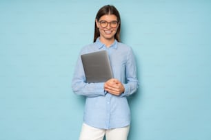 Studio image of cute Caucasian career consultant standing over blue background with gray laptop in hands, smiling at camera in big stylish glasses and casual clothes