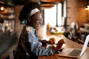 Happy African tourist female visiting new country on her summer vacation. Young Traveller having snack in burger bar, while using laptop to find out new location to reach.