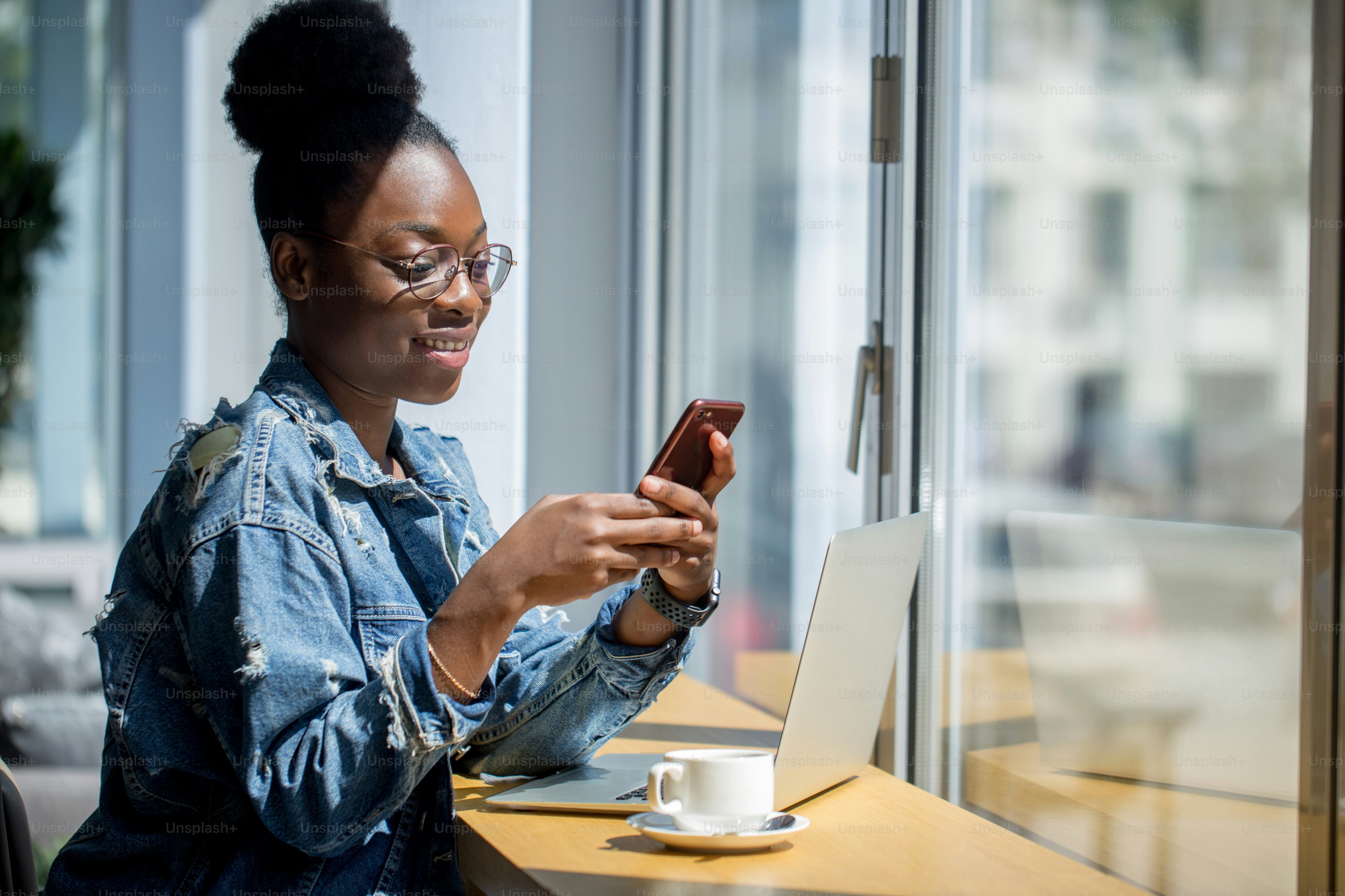 Wanita hipster berkulit gelap dengan gaya rambut Afro berbicara di ponsel pintar di kafe. Wanita muda duduk di meja dengan secangkir kopi dan ponsel berselancar di internet.
