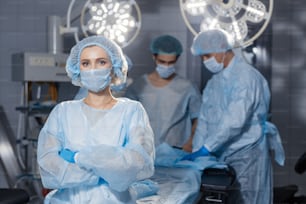 Retrato de cirujana concentrada con uniforme quirúrgico y máscara en quirófano. Joven doctora en el hospital posando con las manos juntas con sus colegas en el fondo.