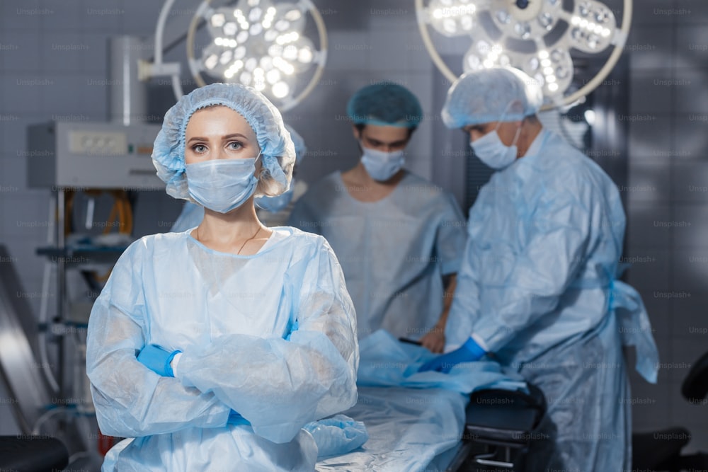 Portrait of concentrated female surgeon in surgical uniform and mask at operating room. Young woman doctor in hospital posing with folded hands with her colleagues on background.