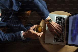 Mãos do velho em elegante terno azul escuro trabalhando no laptop enquanto relaxa no restaurante enquanto carrega vidro de wisky, vista superior