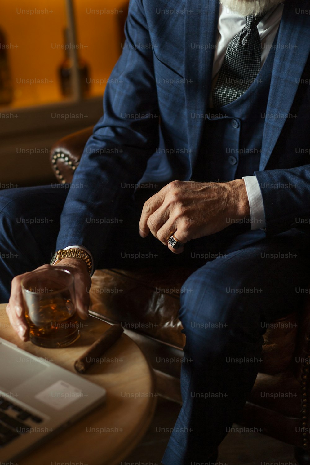 옷을 입은 자신감 있고 부유한 노인은 집에서 노트북 컴퓨터로 일하며 값비싼 술을 마시는 캐비닛이 있는 개인 남성 클럽에 앉아 있습니다.