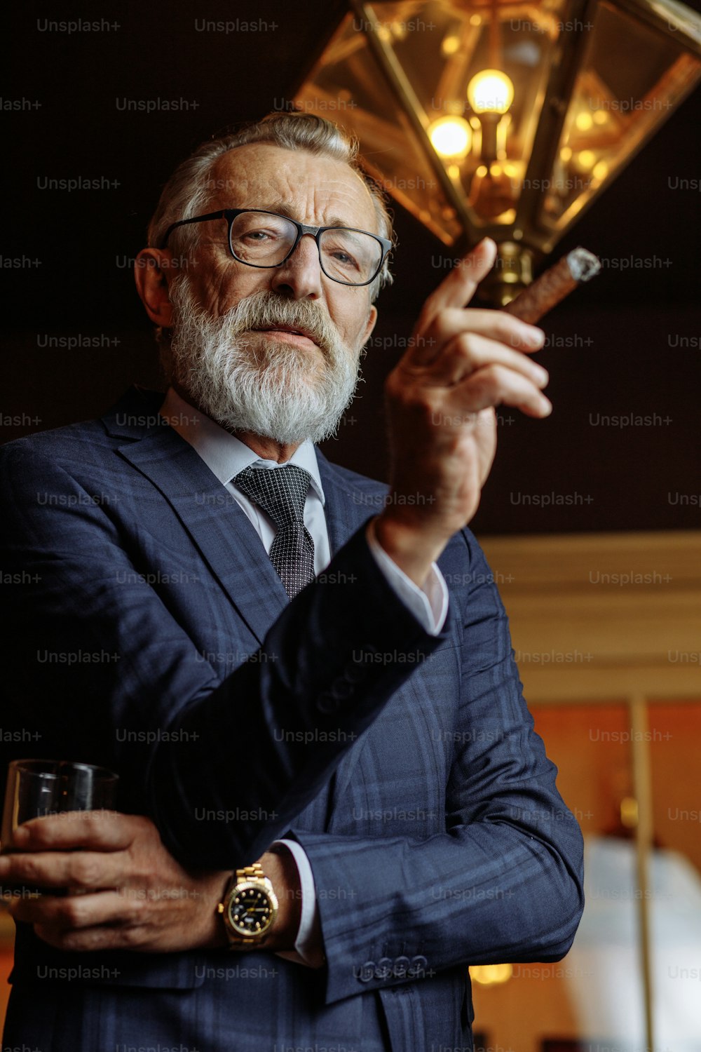 Elderly bearded man dressed in formal clothes, smoking cigar with thoughtful look, tries to concentrate and get idea, looks pensively and seriously at camera, posing in living luxury interior.