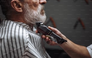 잘 생긴 수염 난 남성 이발사는 이발소에서 잘 생긴 노인을 위해 수염을 모델링합니다. 그는 전기 면도기를 가진 백발의 남자의 수염 디자인을 만들고 있습니다