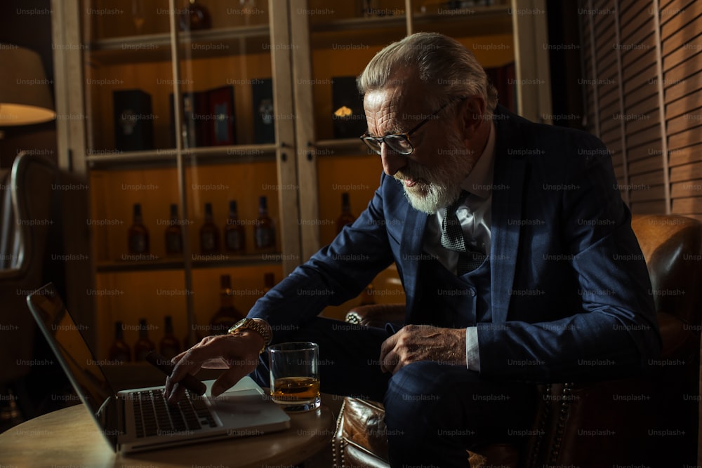 Bearded old-aged playwriter looking at laptop and wrting his new novel while sitting in restaurant at night. To help his creative impulse flow he smoking a cigar and sipping whiskey