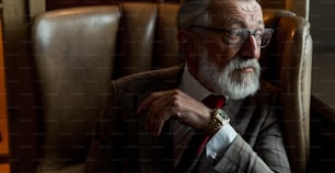 Portrait of trendy fashionable old man in formal suit and glasses, wearing expensive wristwatch, looks at the window while rests in leather chair indoor, close up