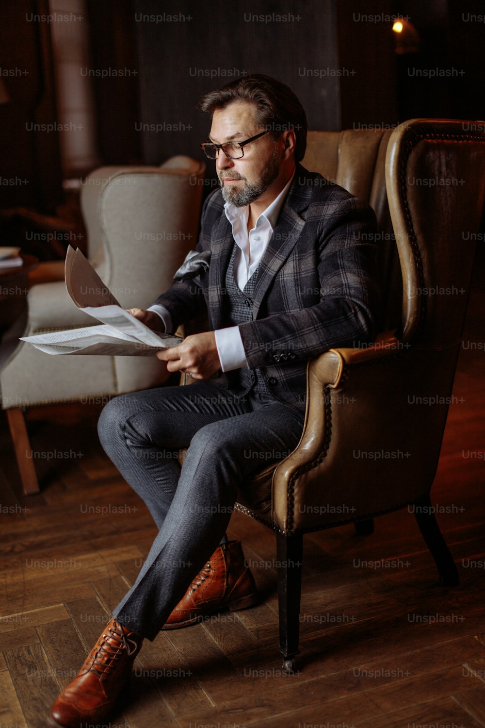 Pensive middle-aged man in elegant custom made suit sitting in leather armchair, reading fresh newspaper in living room with luxury interior.