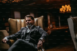 Mature confident businessman in formal bespoke suit looking at camera with serious pensive expression sitting in dark restaurant with artificial lighting and luxurious interior.