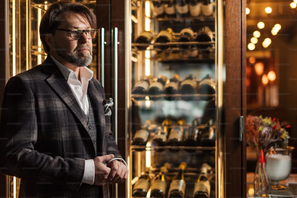 Portrait of a handsome mature male sommelier in tailored suit posing in front of the shelves with bottles at the luxury supermarket or restaurant