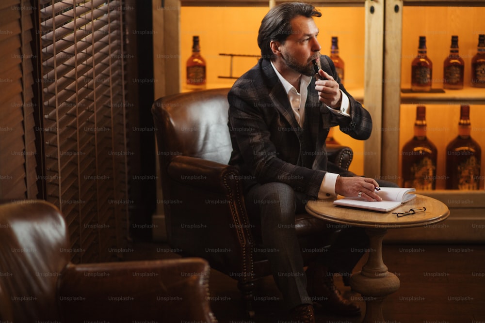 Portrait of busy and hard-working fashion designer smoking pipe and making notes into his diary at his laxurious office.