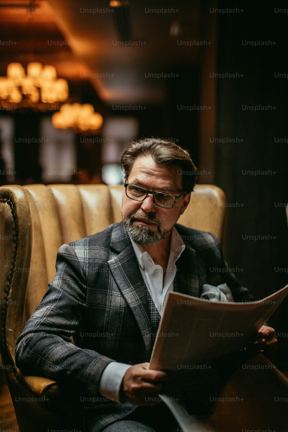 Handsome middle-aged prosperous businessman sitting in leather arm-chair in hotel lobby, wears optical spectacles is reading newspaper while waiting for his room be ready