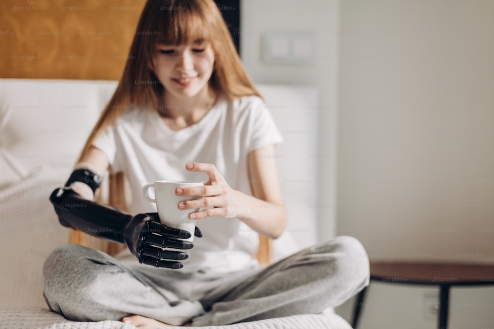 smiling girl with prosthetic cyber arm holding a cup in the room. close up photo.focus on an artificial arm. new life . happiness