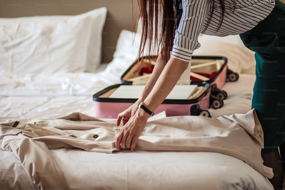 Business woman with long ginger hair, dressed in shirt and pencil skirt packing his shirts in pink suitcase, lying on bed, while staying in hotel
