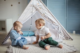 Two gorgeous infant boys of the same age sit on carpet in teepee tent, have fun and play with hristmas garlandin a cozy nursery.