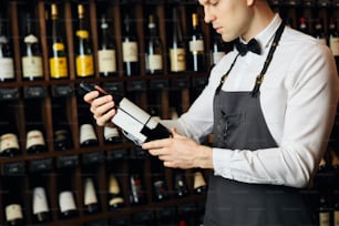 Young caucasian cavist dressed in white shirt and bowtie working in big vine shop presenting a bottle of red wine to customer