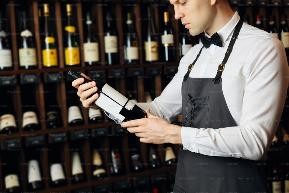 Junger kaukasischer Kavist in weißem Hemd und Fliege, der in einem großen Weinladen arbeitet und dem Kunden eine Flasche Rotwein überreicht