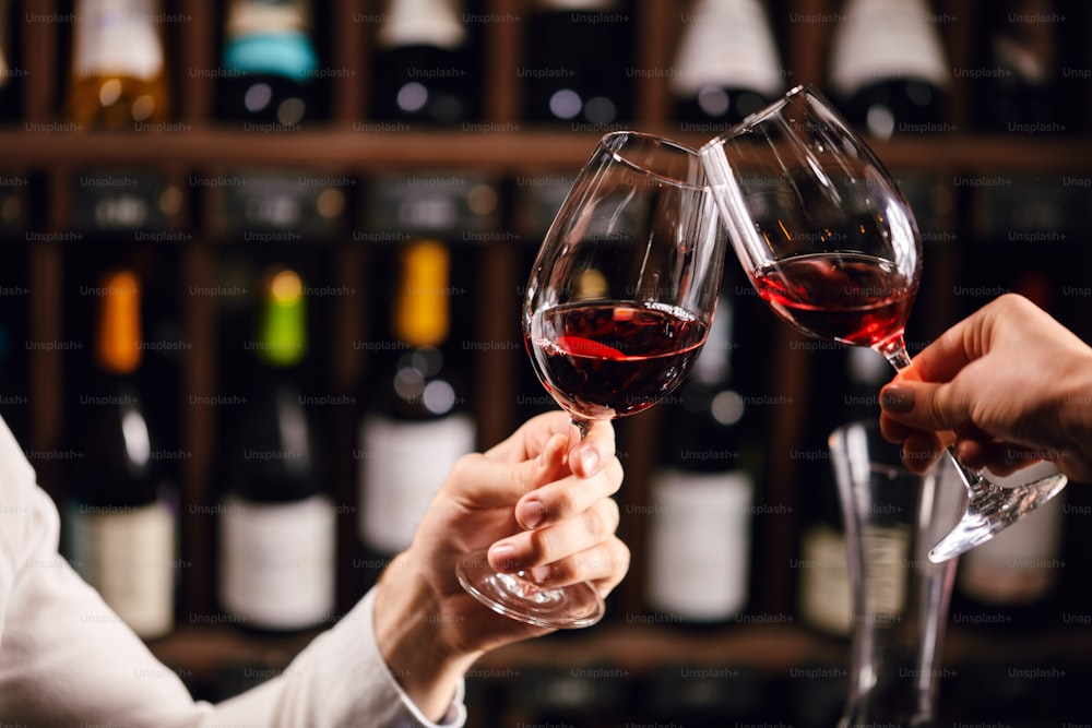 Dos personas tintineando con copas de vino tinto, celebrando el éxito o brindando en un restaurante de vinos, contra estantes con botellas de vino, de cerca.