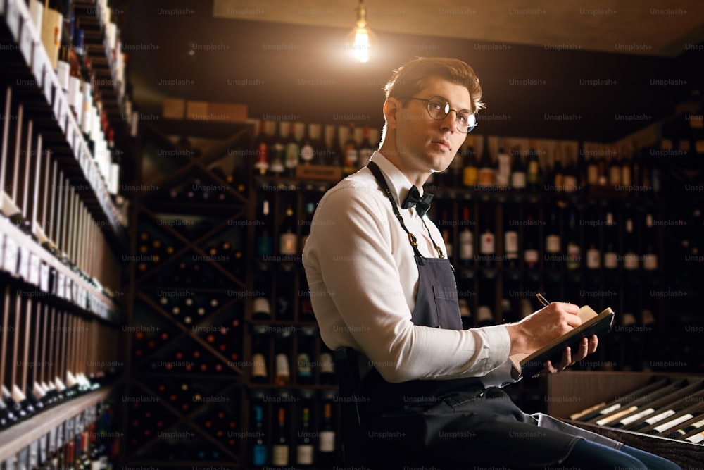 Cavista profissional masculino examina as garrafas com vinho na loja de vinhos, segurando amostra maravilhosa, pronto para falar sobre este vinho para os clientes