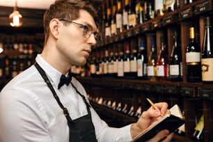 Professioneller männlicher Sommelier in Fliege und Schürze schreibt auf Notizblock und notiert Informationen über Weinflaschen im Weinhaus