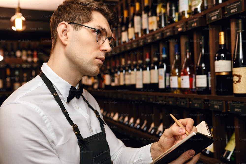 Professional male Sommelier dressed in bowtie and apron writes at notepad, putting down information on wine bottles in wine house