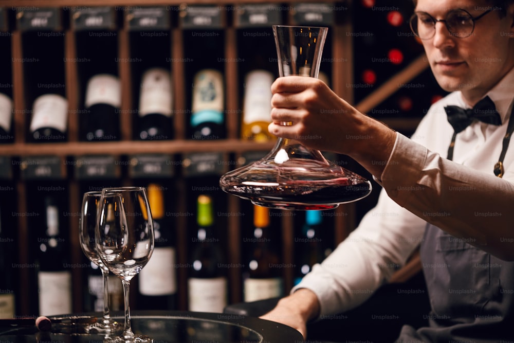 Catador o degustador de vinos mezclando vino tinto en jarra para hacer un color perfecto en el fondo con estantes de botellas de vino en la casa de vinos.
