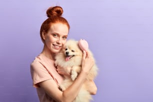 charming ginger girl with toothy smile using brush to take care of her adorable pet. close up portrait, girl groomer preparing a dog for a party