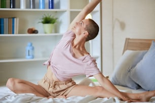 Mujer caucásica en la cama practica ejercicios de yoga, habitación blanca de fondo. Manos levantadas.