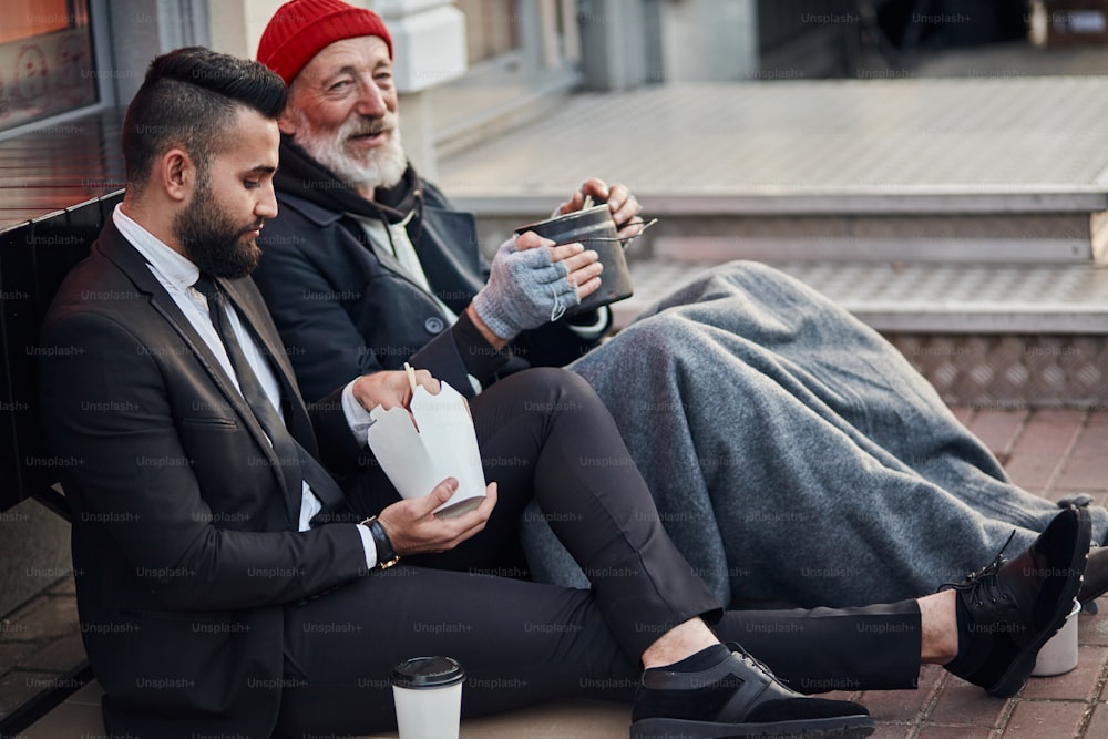 Hübscher Geschäftsmann im Anzug, der mit einem Obdachlosen zusammen auf dem Boden sitzt, hört sich seine Lebensgeschichte an. Kontrastieren Sie Menschen, Reiche und Arme, aber das spielt keine Rolle