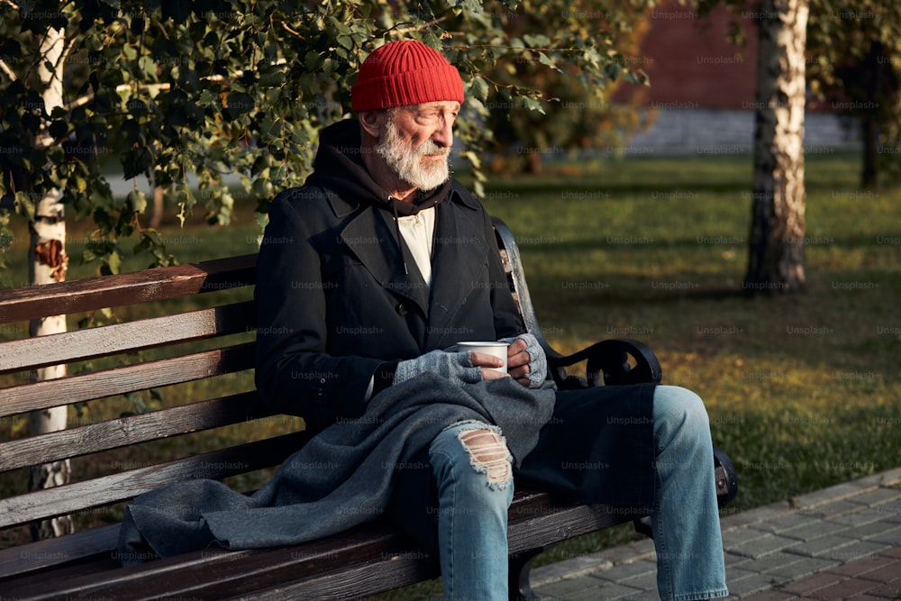 Älterer Bettler, der verzweifelt auf die leere Straße schaut und sich auf Geld freut, Hilfe. Gekleidet in roten Hut und warmem Mantel