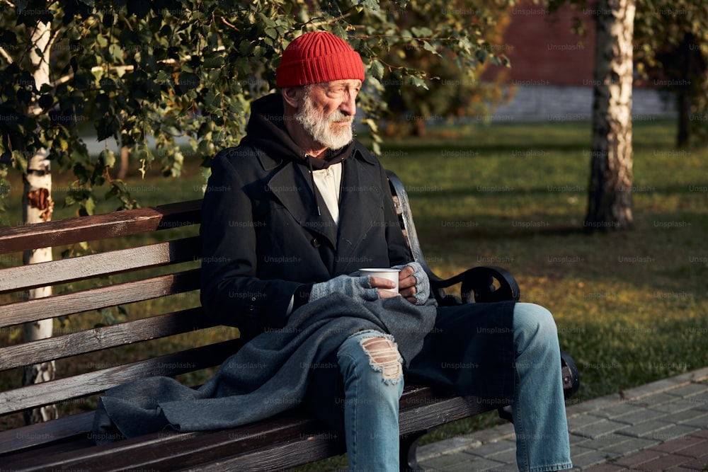 Senior beggar male desperately looking at empty street, looking forward for money, help. Dressed in red hat and warm coat