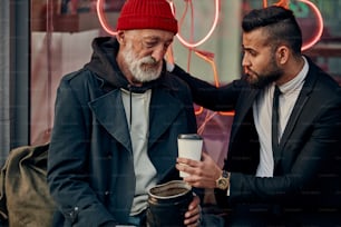 L'uomo d'affari in giacca offre caffè al mendicante maschio in strada che indossa vestiti sporchi casual
