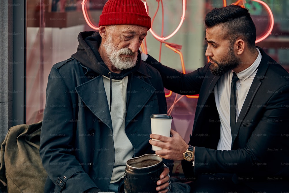 Business man in suit offers coffee to beggar male in street wearing casual dirty clothes