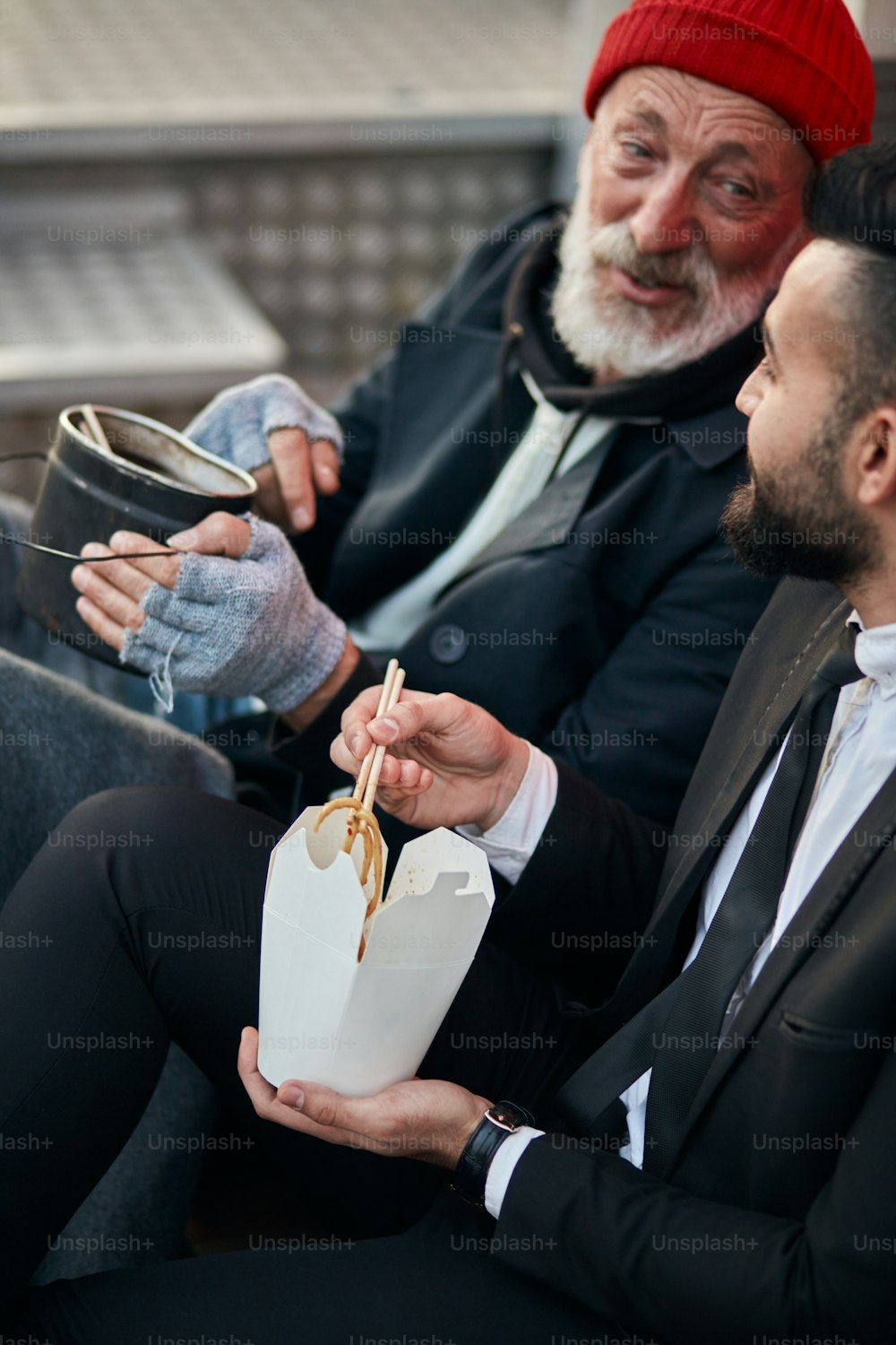 Beggar man with kind eyes, happily look at man. Handsome businessman in suit sitting on floor with homeless man together, listen to story of life. Contrast people, rich and poor, but doesn't matter
